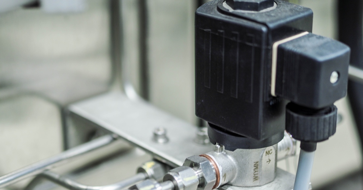 A close-up of a black high-pressure solenoid valve connected to a metal pipe that’s part of an industrial system.