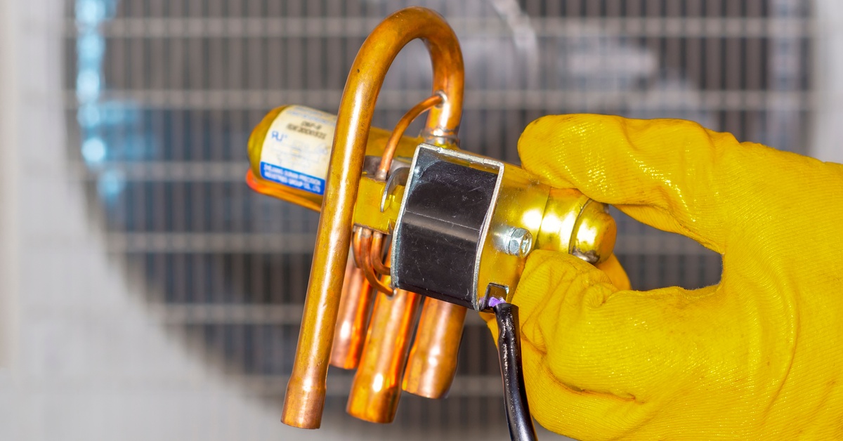 A hand in a yellow rubber glove holds one end of a four-way solenoid valve, with a blurred HVAC system in the background.