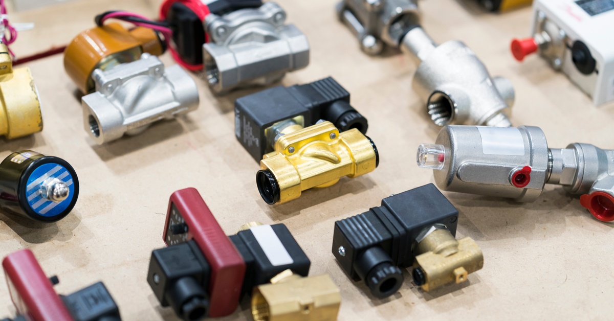 Eight different industrial solenoid valves arranged beside one another sit on a wooden table by slightly blurred equipment.