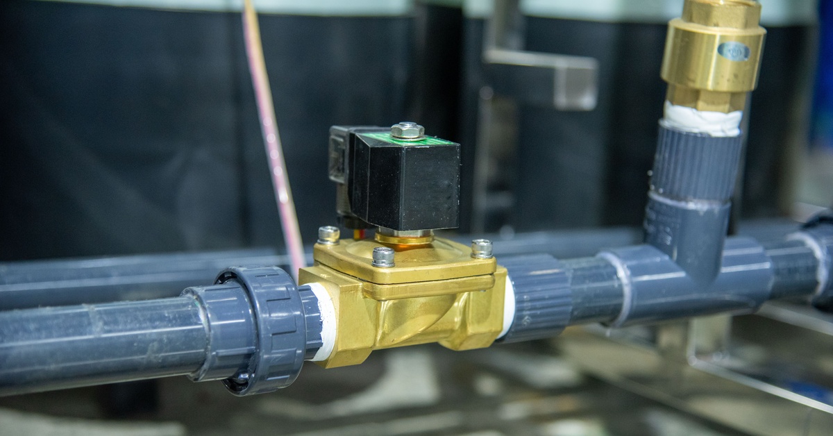 A brass solenoid valve connected to a gray, industrial pipe system inside of a building; the background is slightly blurred.