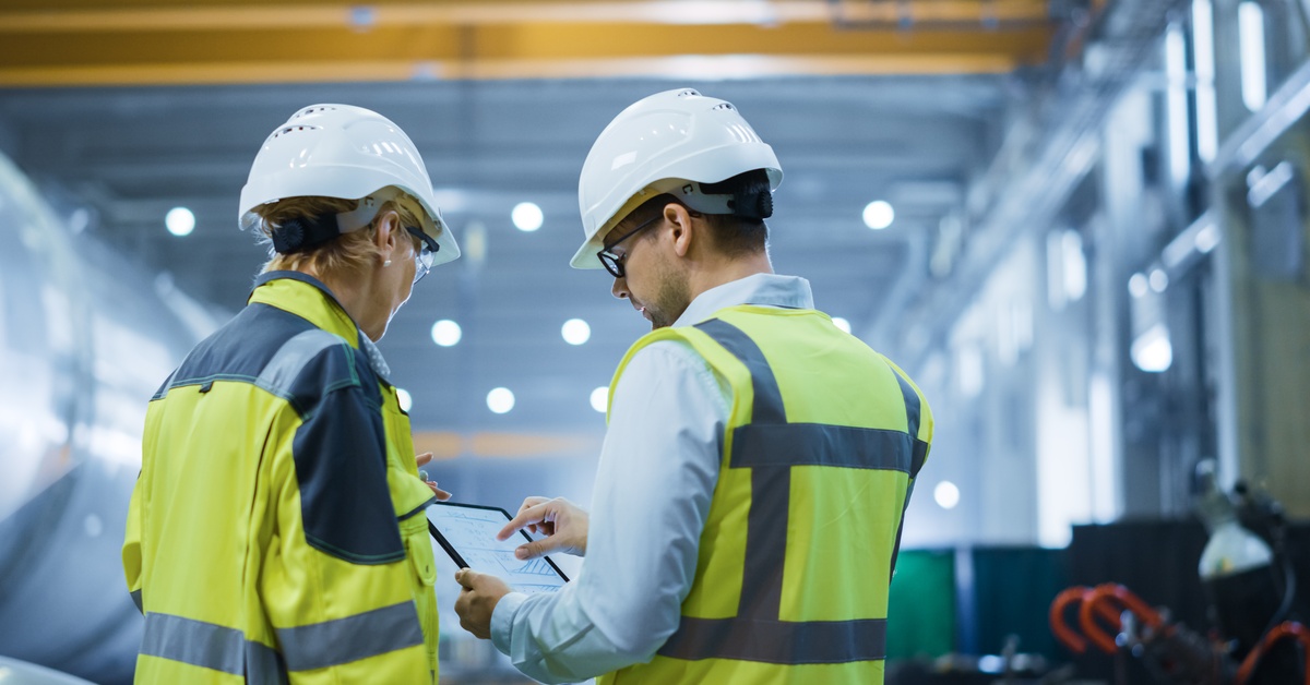 Two people in reflective gear and hard hats stand beside one another, looking at a tablet screen that one of them holds.