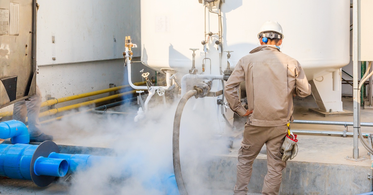 A technician in a hard hat operates a liquid nitrogen storage tank in a factory. White vapor surrounds the equipment.