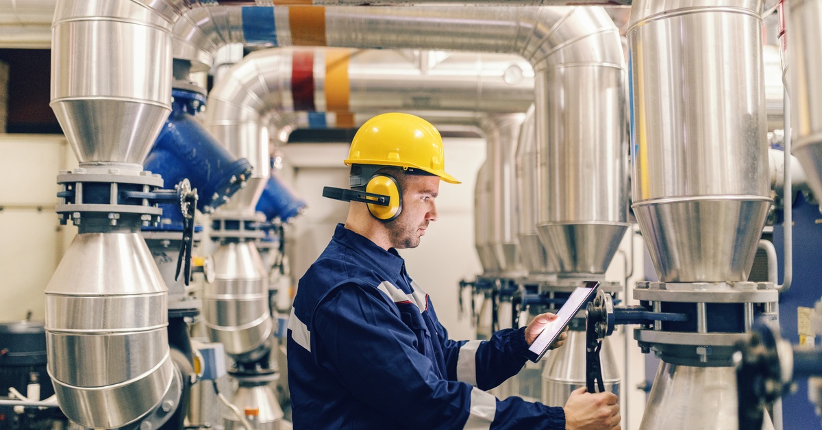 A man in a yellow hard hat, soundproof headphones, and a safety jacket holds a wrench against a valve.