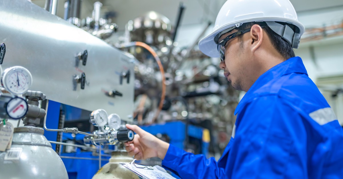 An engineer in a blue work jacket and white hard hat holds a clipboard and inspects a valve in a factory.