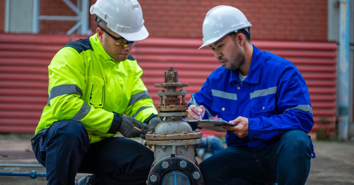 Two workers in safety gear inspect a large pipe with valves in an industrial setting; one worker takes notes on a clipboard.