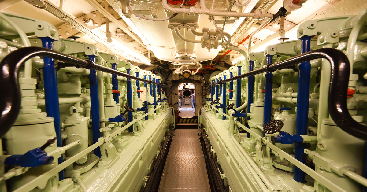 A submarine's diesel engine compartment with green machinery, pipes, and a narrow walkway leading to a circular opening.