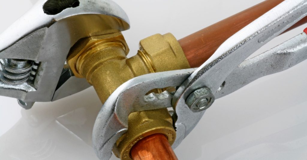 Two wrenches compress the sides of a gold three-way valve connected to copper pipes, set against a white background.