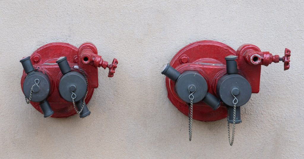 A pair of red and gray 2-way valve hose connectors beside one another, mounted to the wall of an unseen building.
