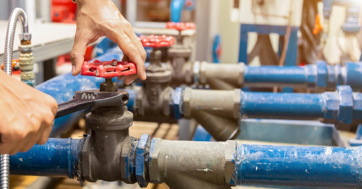 An unseen person uses a wrench to repair blue industrial water pipes with red valves in an indoor setting.