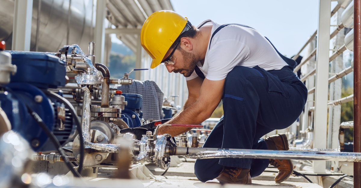 A worker in a yellow helmet and blue overalls kneels beside a pipe system outside and works on one of the valves.