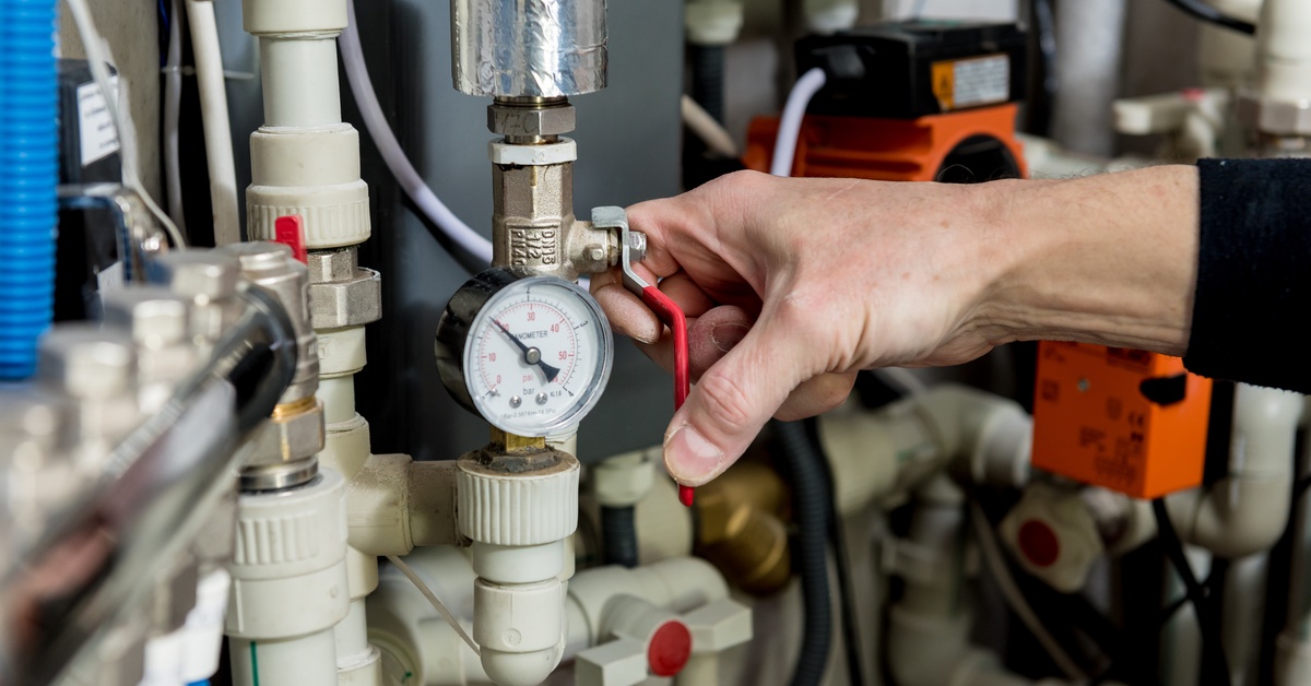 A person’s hand adjusts a red valve on a modern heating system with pipes and gauges in a boiler room.