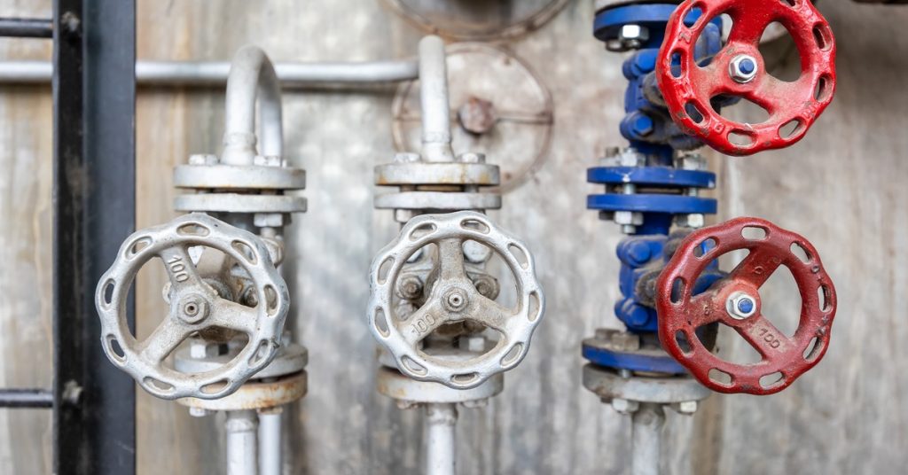Industrial manual hydraulic valves with spoked handwheels, some painted red and blue, in a factory setting.