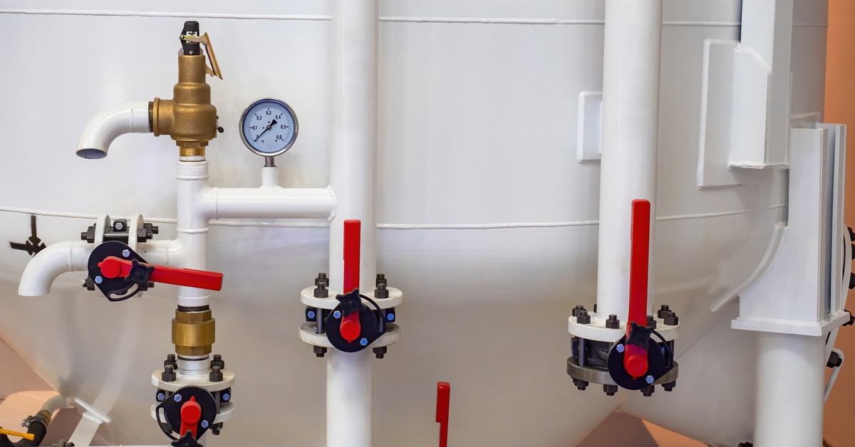 White industrial pipes with brass valves and pressure gauges on a large cylindrical tank in an industrial setting.
