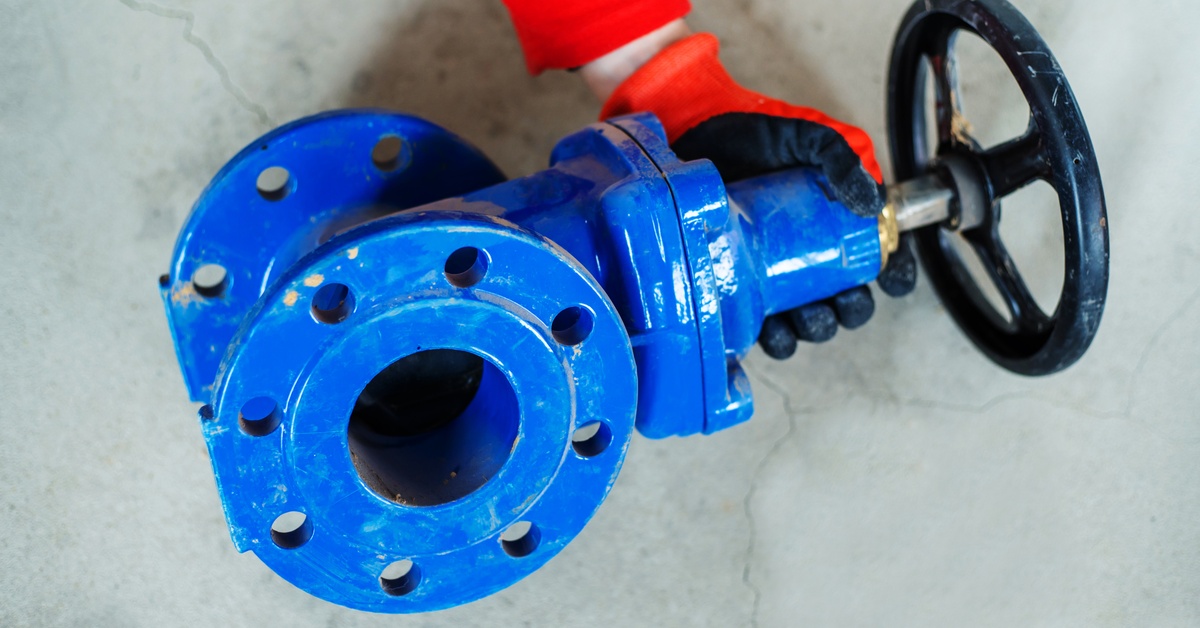 A close-up of a blue water valve with a black handwheel. A person wearing a black and red glove holds the valve.