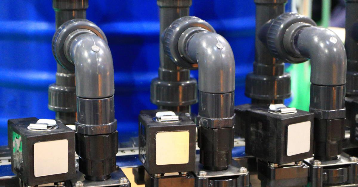 Electro-hydraulic valves with gray pipes and black rectangular components, set against a blurred industrial background.