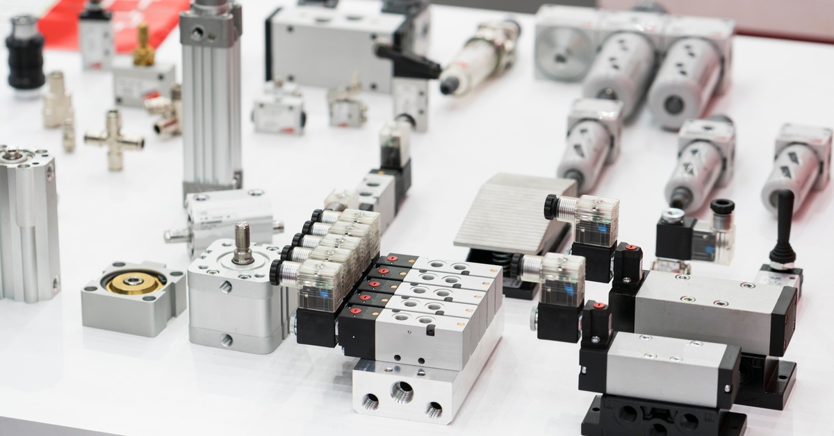 A collection of industrial solenoid valves, pneumatic cylinders, and metallic components arranged on a white surface.