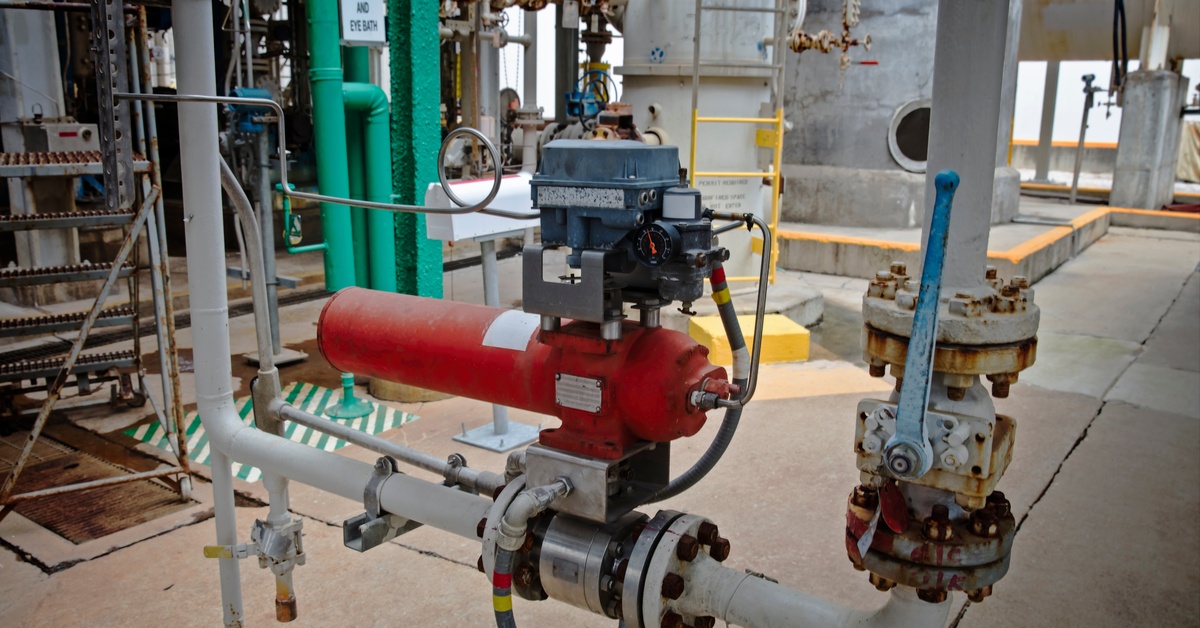 A red pneumatic solenoid shutoff valve connects to white pipes within an industrial facility with concrete floors.
