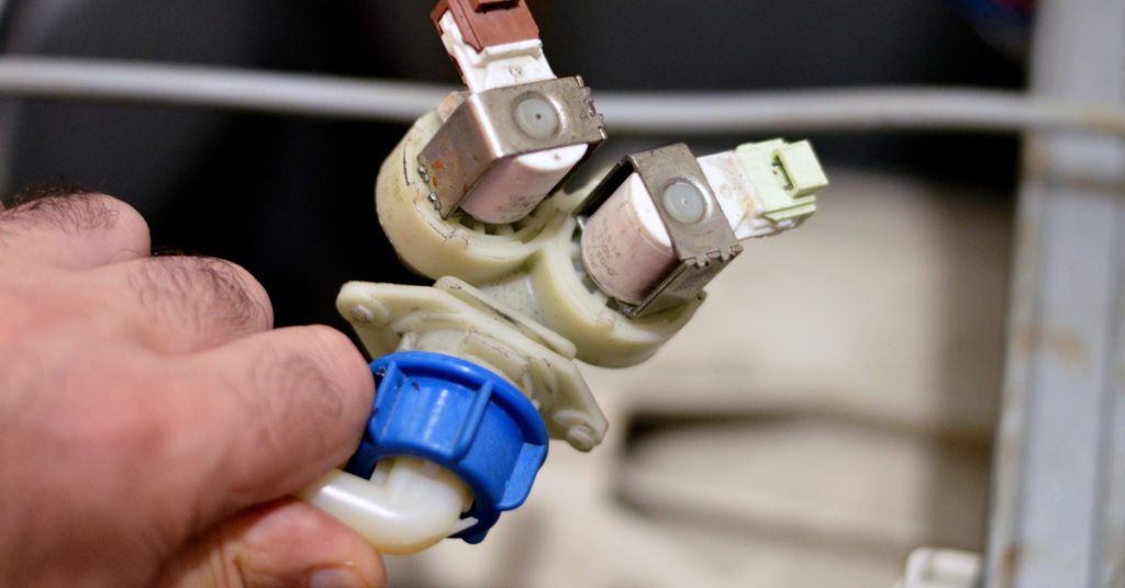 A hand holds a plastic inlet water valve with attached connectors and wiring inside a washing machine compartment.