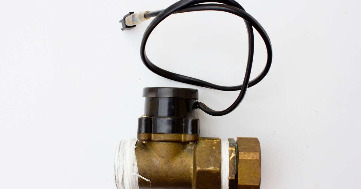 A solenoid valve with an attached cable rests on a white surface, with its threaded metal ends and wiring clearly visible.