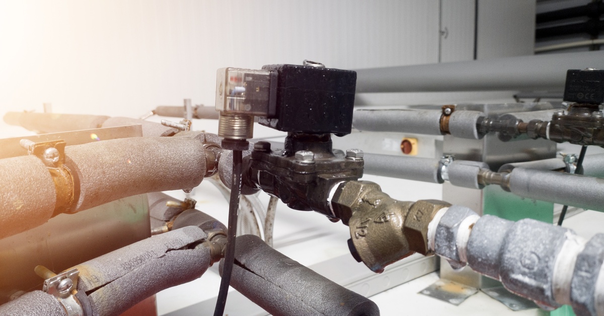 A solenoid valve with a black coil connected to insulated pipes and metal fittings inside industrial equipment.