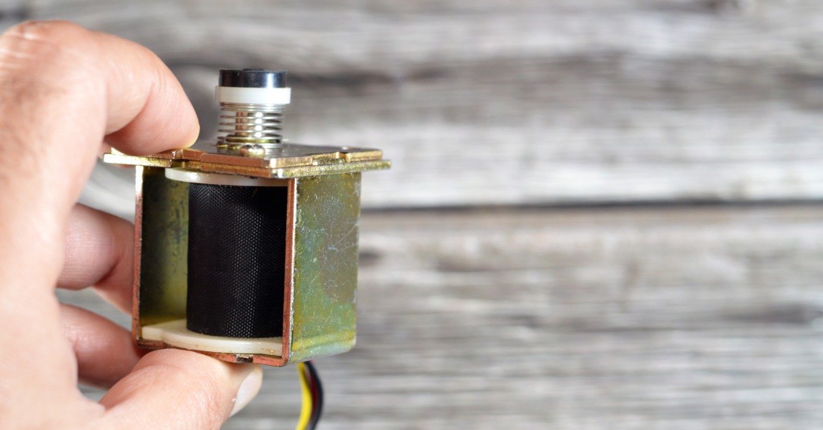 A hand holding a small metal solenoid valve with a black coil against a blurred wooden board background.