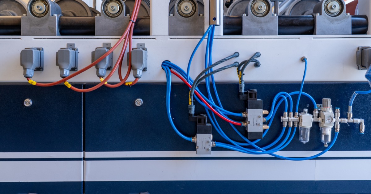 An industrial machine panel with rollers, pneumatic valves, blue tubes, red cables, and mounted connectors.