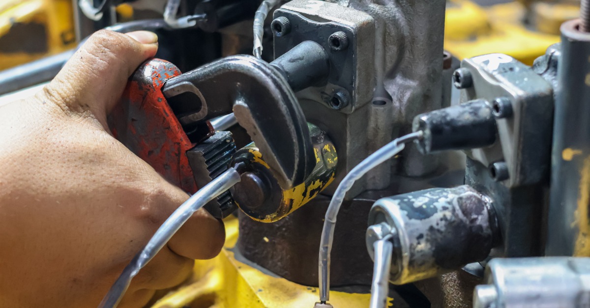 A hand holding a red pipe wrench around a black metal fitting among hoses, valves, and yellow machinery parts.
