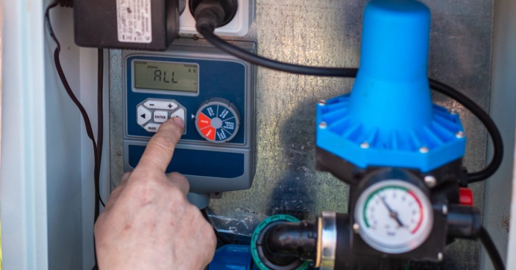 A hand pressing a button on a blue digital pump controller with a round pressure gauge and connected pipes.