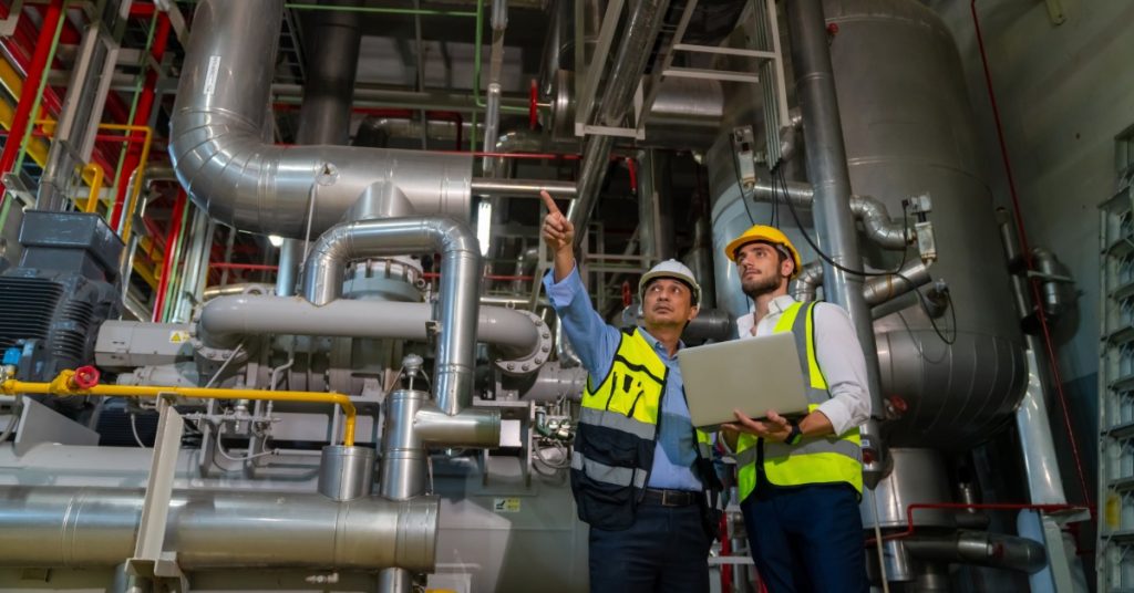 Two workers in hard hats using a laptop beside electrical control panels in an industrial control room.