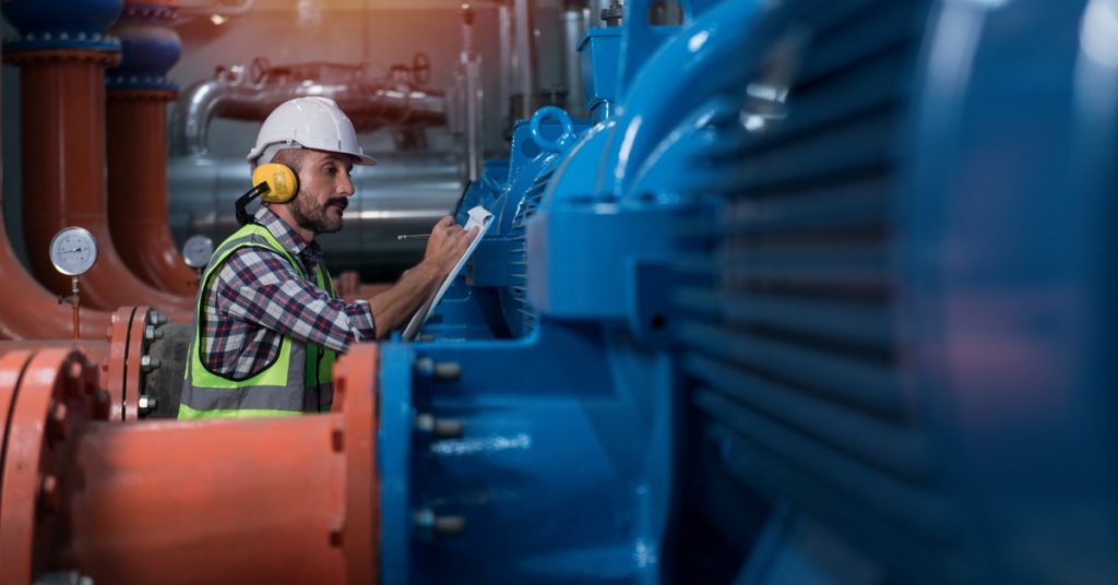 A man in a hard hat and yellow hearing protection writes on a clipboard beside blue pumps and orange pipes.