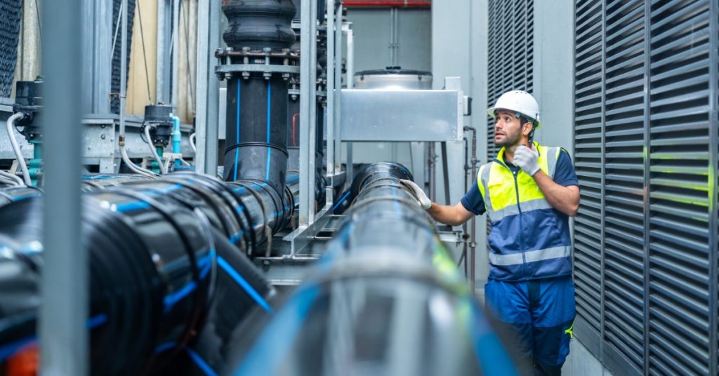 A worker wearing a white hard hat and safety vest is using a radio beside large black pipes and metal panels.