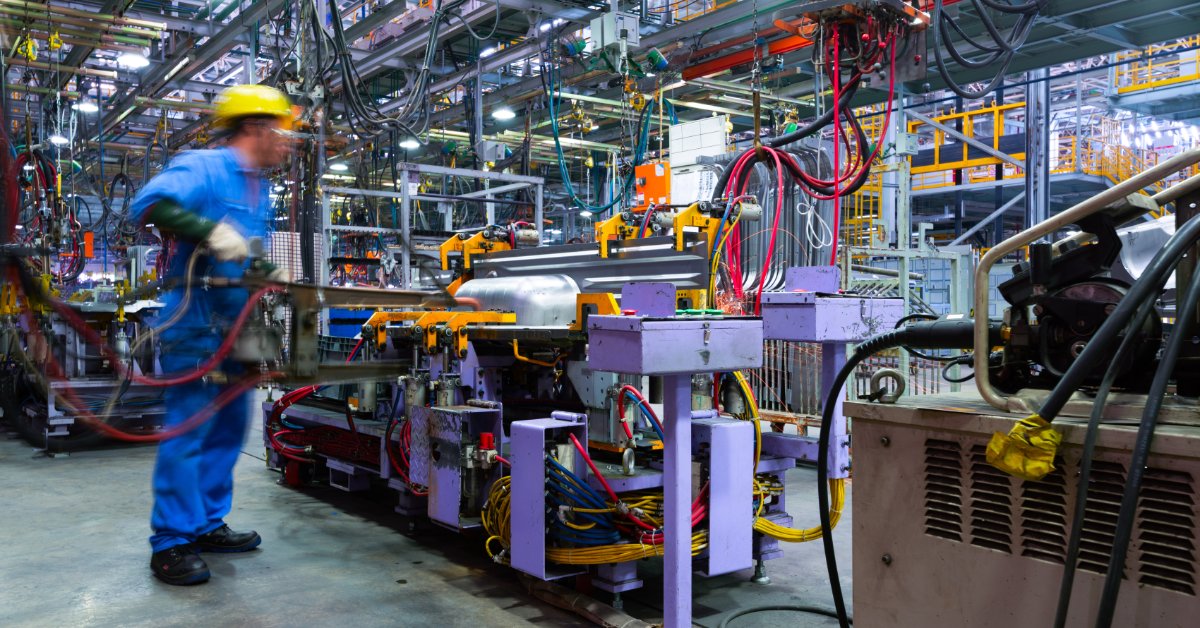 A worker approaches a workstation at an automated factory line with overhead machinery and bright industrial lighting.