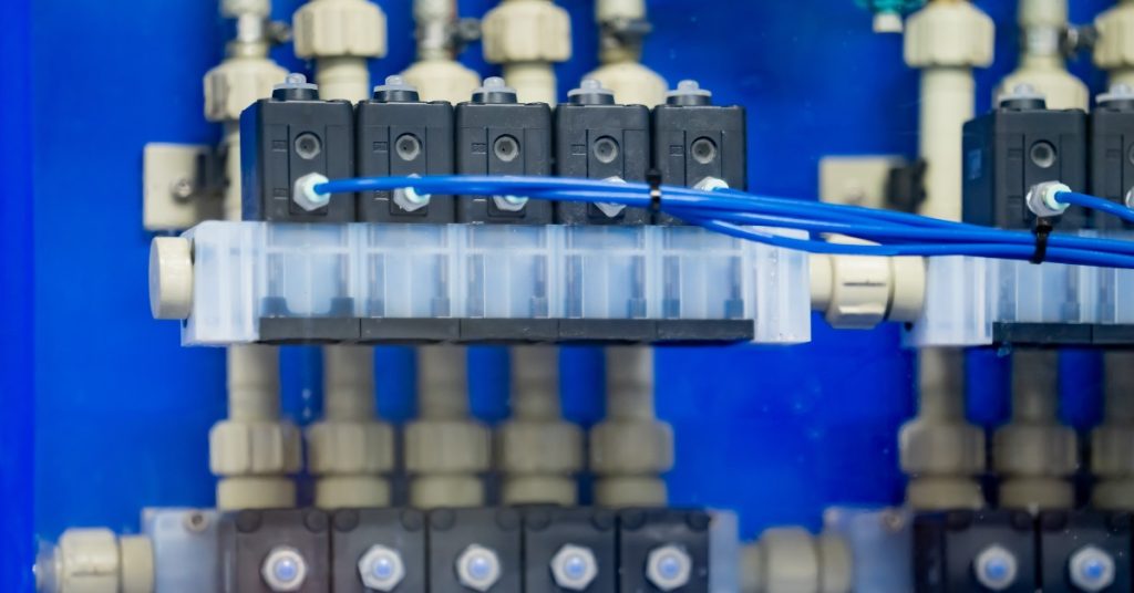 A solenoid valve assembly connected to plastic pipe fittings and blue control lines inside an industrial panel.
